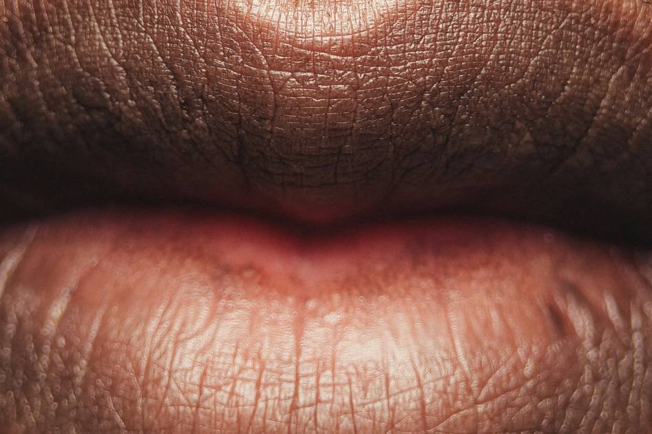 A close-up of glossy, plump lips with a lip plumper gloss tube in the background on a soft pink surface